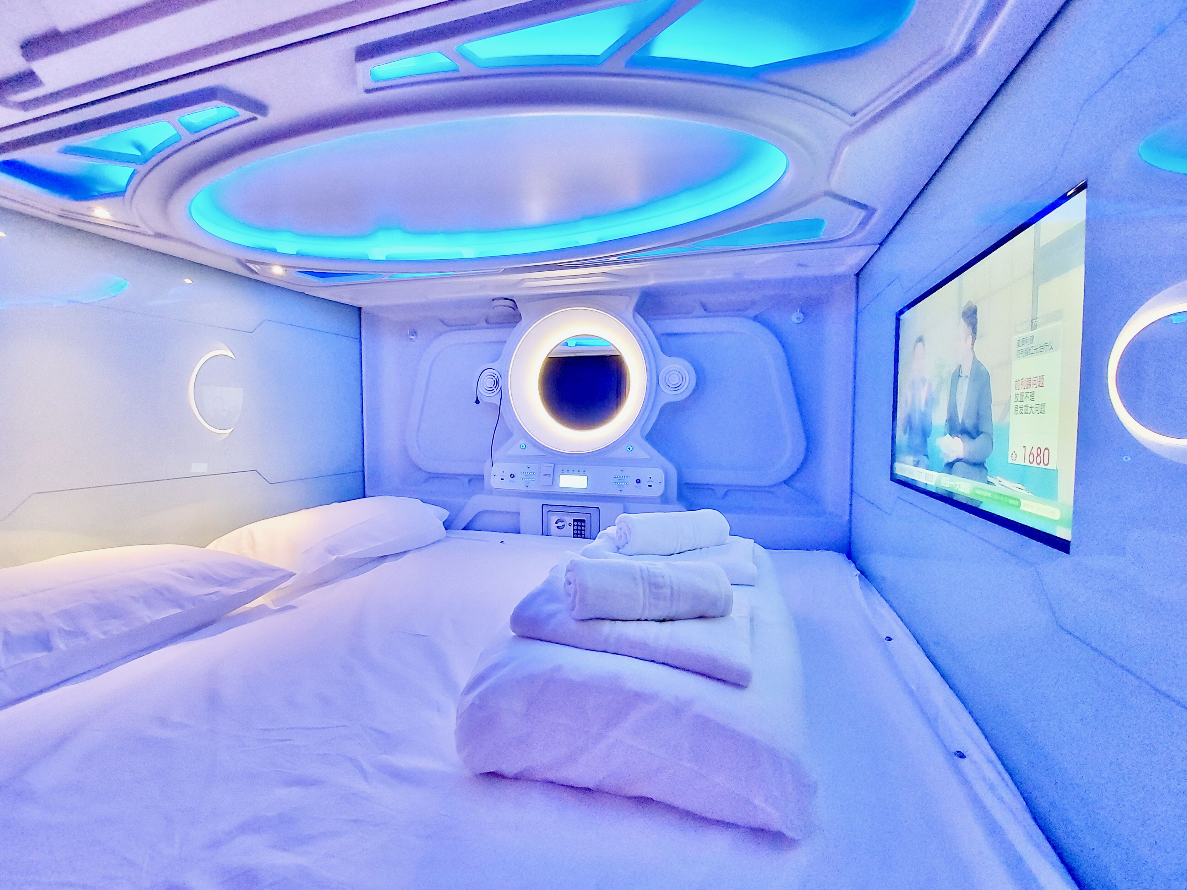 A modern room with a bed, a massage chair, and a wall-mounted screen displaying information.