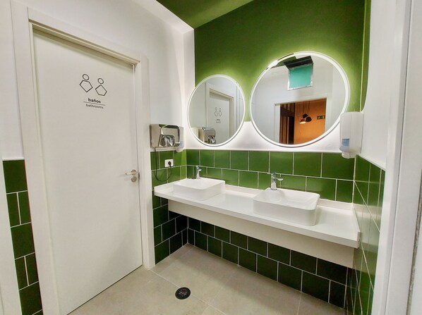Un baño con dos lavamanos, paredes con azulejos verdes, una puerta blanca y un espejo con marco redondo.