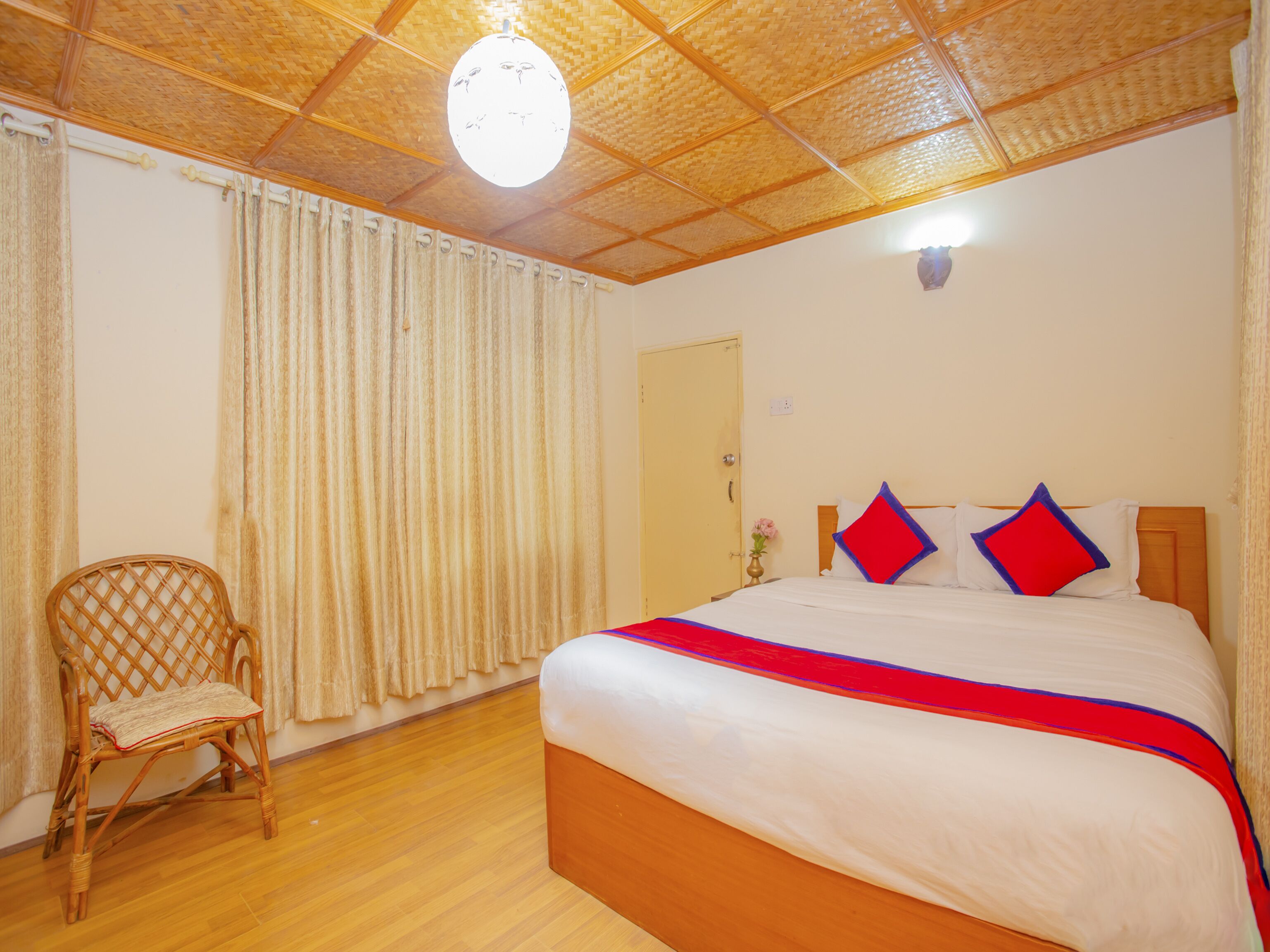A hotel room with a wooden ceiling, a bed with a red and blue bedspread, a wicker chair, and a wooden floor.