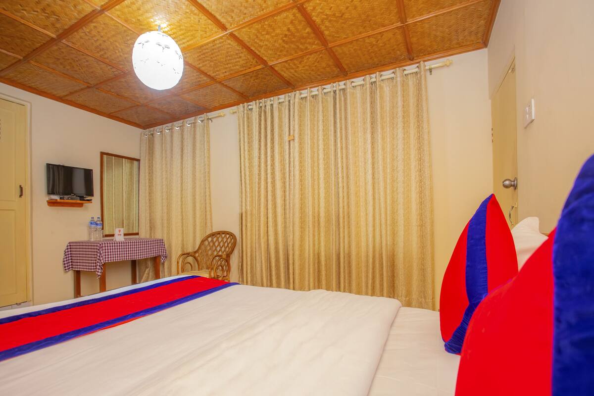 A hotel room with a large bed, a television on a shelf, a chair, and a table with a checkered tablecloth.