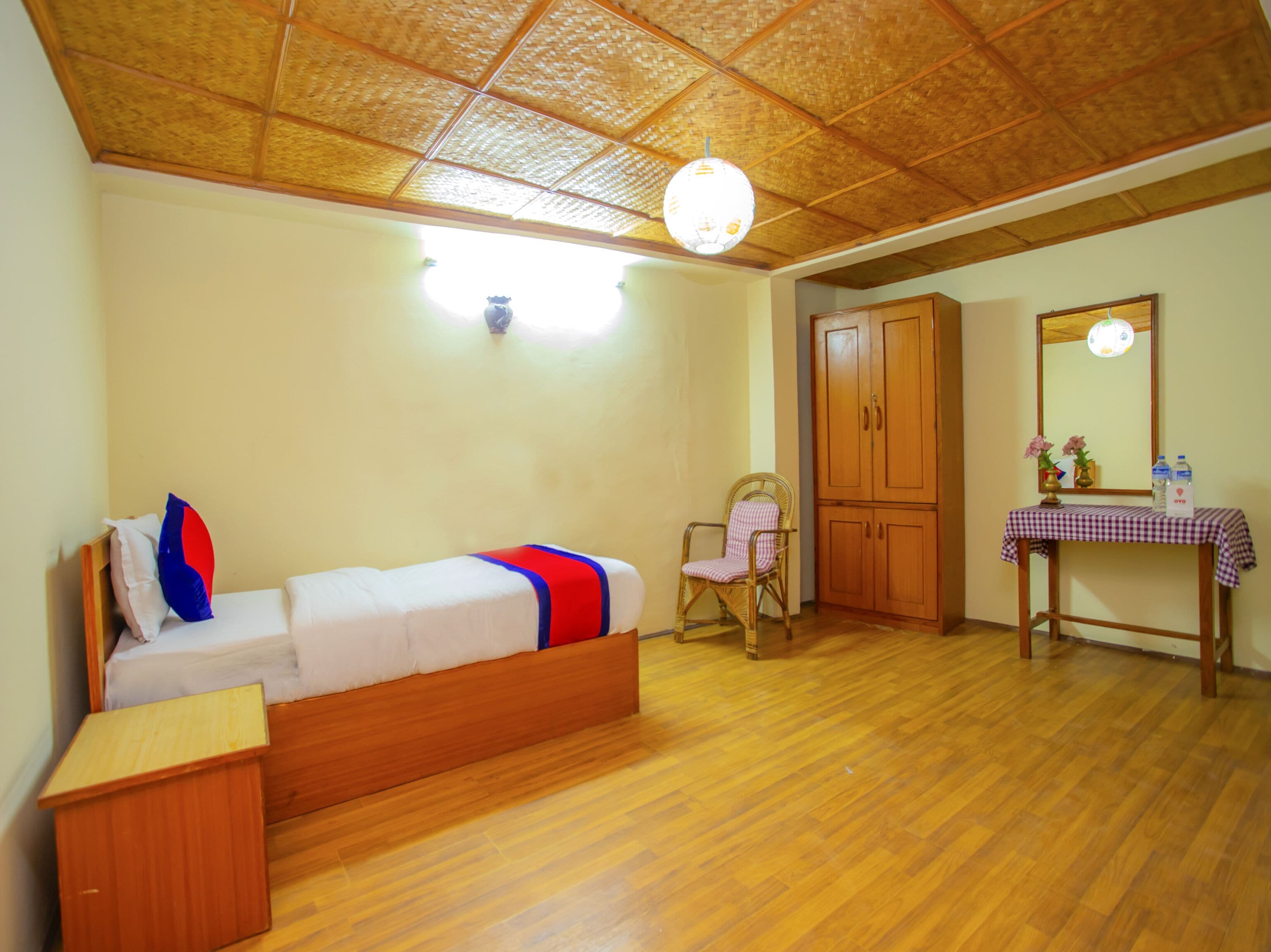 A room with a wooden ceiling, a bed with a red and blue blanket, a chair, a wooden wardrobe, a table with a checkered tablecloth, and a mirror.