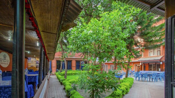 A courtyard with a central tree, brick pathway, and outdoor seating area with blue tables and chairs. - Sweet Home Hotel (Bhaktapur)