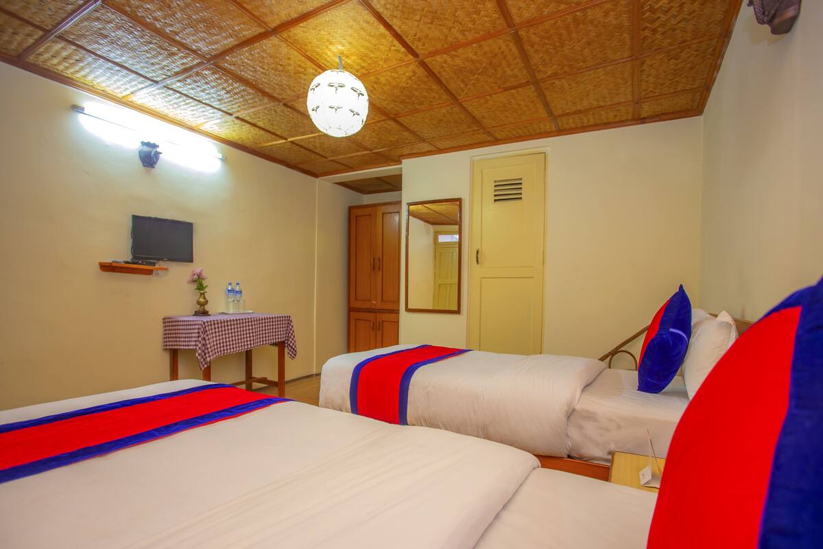 A hotel room with two beds, a small table with a checkered tablecloth, a television on a shelf, and a wooden ceiling.