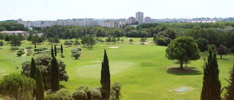 A golf course with a city skyline in the background, surrounded by trees and a building with outdoor seating.