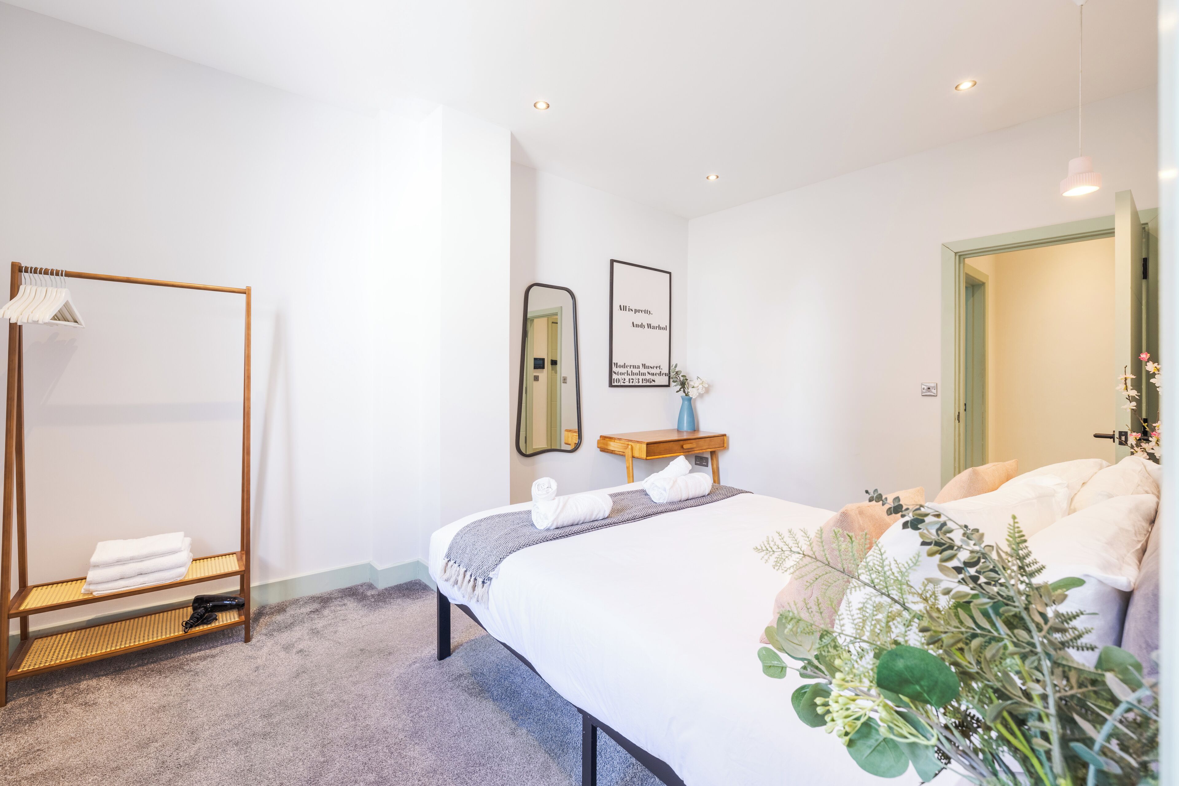 A hotel room with a bed, a wooden wardrobe, a mirror, a framed quote on the wall, and a small table with a vase of flowers.