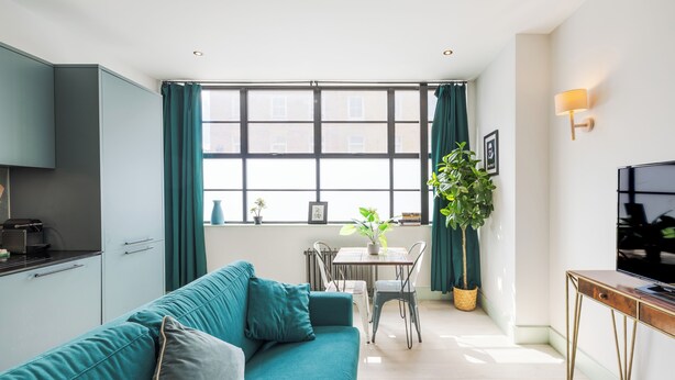 Una sala de estar moderna con un sofá color turquesa, una mesa de comedor con sillas, un televisor y un ventanal con cortinas.