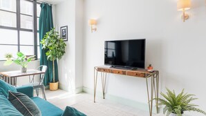 Una sala moderna con sofás turquesa, un televisor de pantalla plana sobre un soporte de madera, una pequeña mesa redonda con una planta y un comedor con mesa y sillas.
