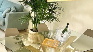 Una sala moderna con un sofá, una mesa baja y una mesa de comedor de vidrio con sillas. Hay una planta en maceta y una botella de champán sobre la mesa.