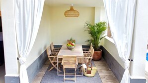 Una mesa de comedor de madera con un frutero, rodeada de sillas, en una habitación con cortinas blancas y una lámpara colgante.