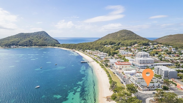 An aerial view of a coastal town with a beach, clear blue waters, and hilly terrain in the background. - Fleetwood, 3/63 Shoal Bay Road - Relaxed living with water views overlooking Shoal Bay (Shoal Bay)