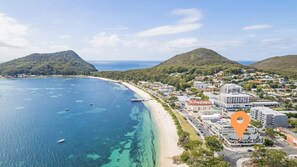 An aerial view of a coastal town with a beach, clear blue waters, and hilly terrain in the background.