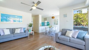 A modern living room with two grey sofas, a dining area with white chairs, a kitchen in the background, and a large window with a view of trees and parked cars. - Fleetwood, 3/63 Shoal Bay Road - Relaxed living with water views overlooking Shoal Bay (Shoal Bay)