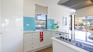 A modern kitchen with a window, a sink, a fire extinguisher sign, and a countertop with a bowl of fruit and glasses. - Fleetwood, 3/63 Shoal Bay Road - Relaxed living with water views overlooking Shoal Bay (Shoal Bay)
