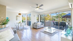 A spacious living area with a grey sofa, a glass coffee table, and a dining table with chairs. There is a ceiling fan and a large window offering a view of the outdoors. - Fleetwood, 3/63 Shoal Bay Road - Relaxed living with water views overlooking Shoal Bay (Shoal Bay)