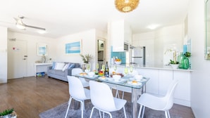 A modern living room with a glass dining table, white chairs, a grey sofa, and a kitchen area with white cabinets and a large mirror. - Fleetwood, 3/63 Shoal Bay Road - Relaxed living with water views overlooking Shoal Bay (Shoal Bay)
