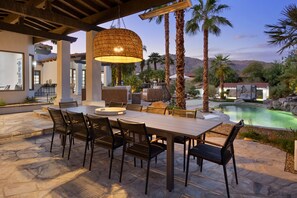 An outdoor dining area with a table and chairs, a hanging light fixture, and a pool in the background.