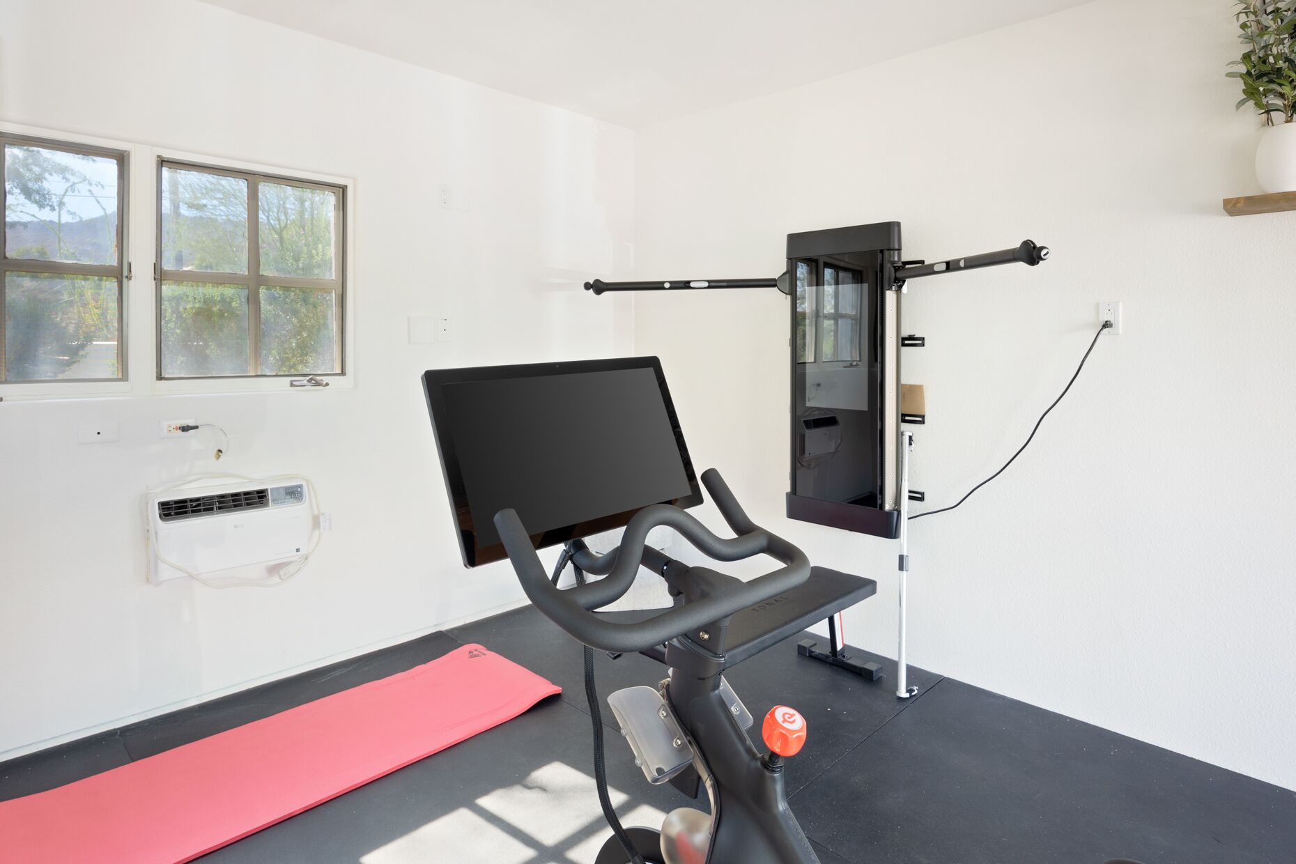 Una habitación con una bicicleta estática, una colchoneta roja, un televisor montado en la pared y una ventana con vistas a árboles.