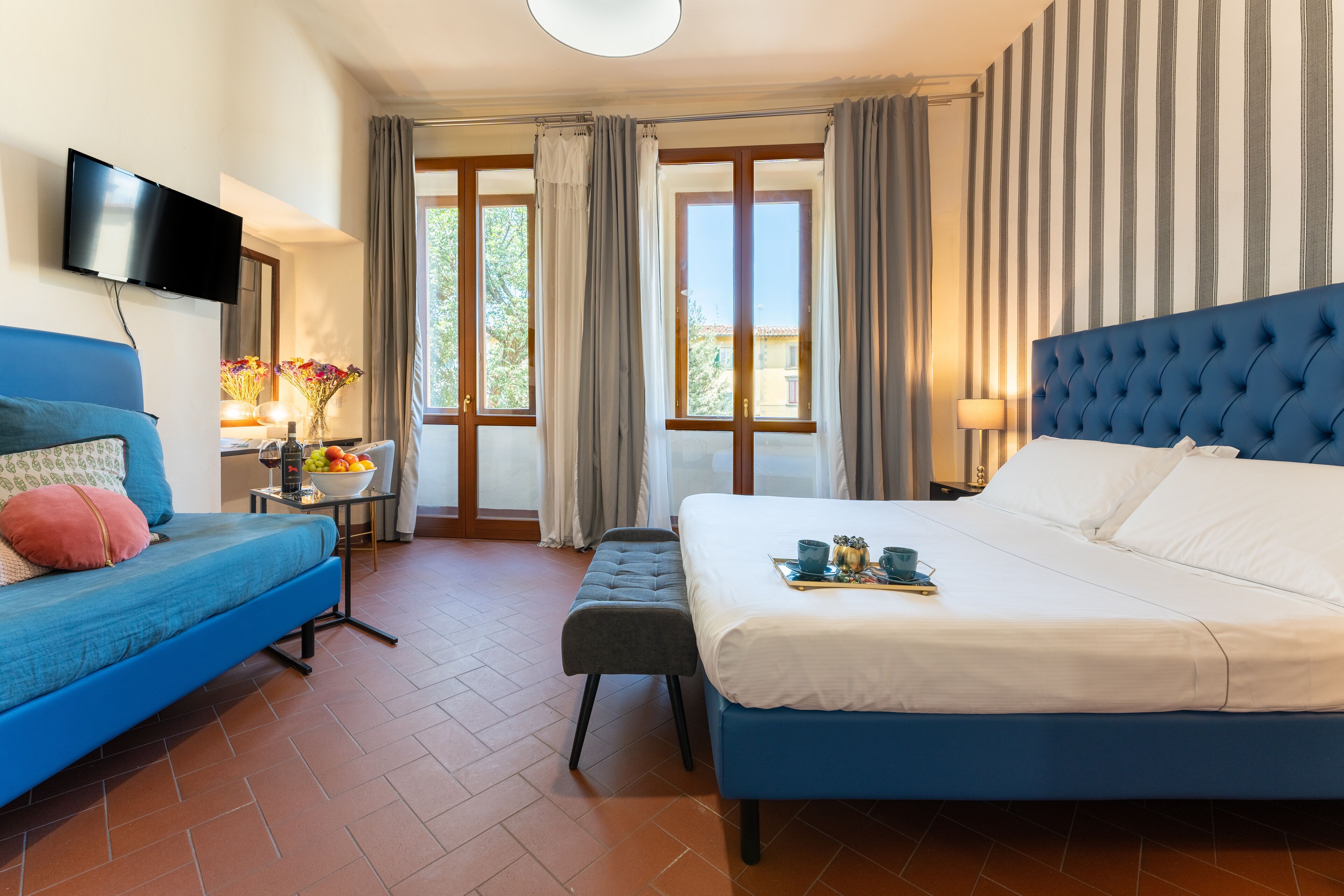 A hotel room with a large bed, a blue sofa, a small table with a lamp, and a TV mounted on the wall.