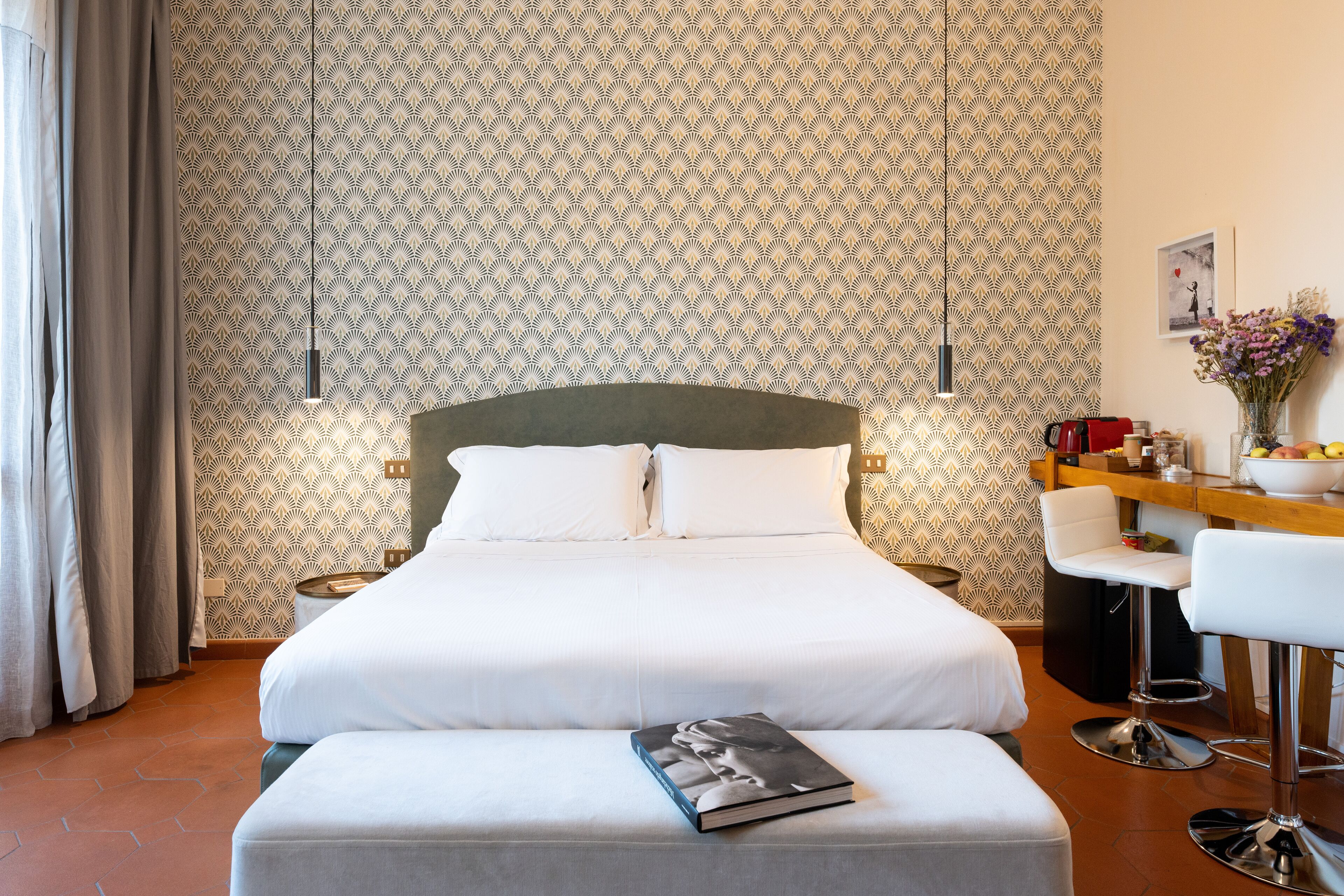 Quarto de hotel bem arrumado, com uma cama grande, mesa de cabeceira, uma cadeira e um pequeno bar.