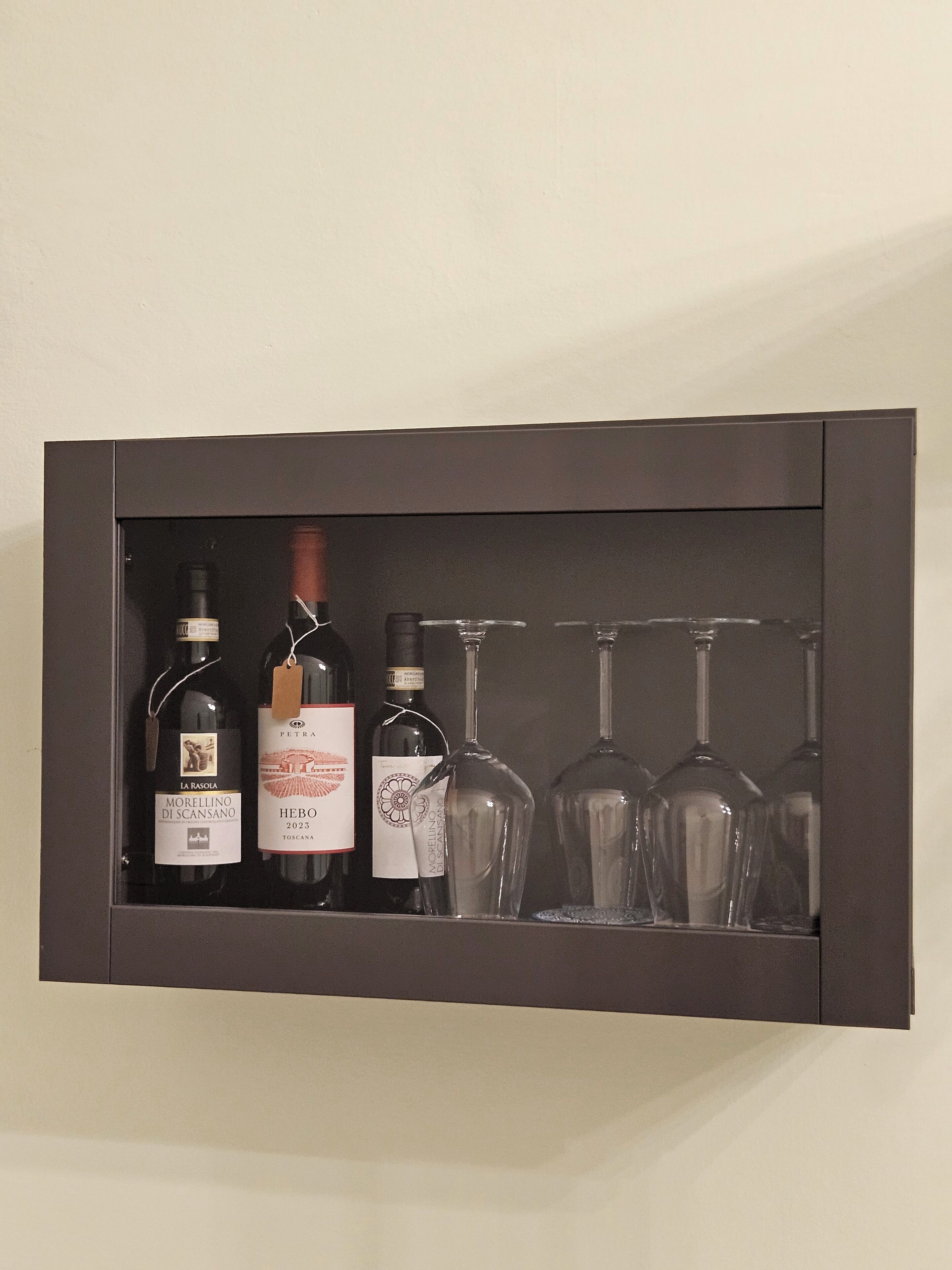 A dark-colored cabinet with three wine bottles and four wine glasses inside.