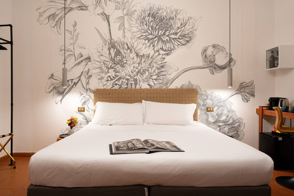 A neatly made bed with a book on it, a nightstand with a vase of flowers, and a wall with a large floral mural.