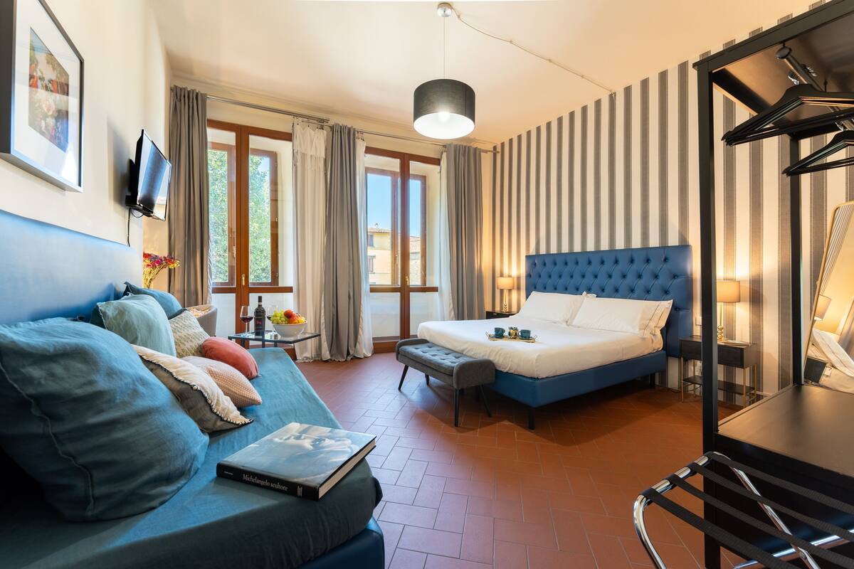 A hotel room with a blue sofa, a bed with a blue headboard, a small table with a lamp, and a TV mounted on the wall.