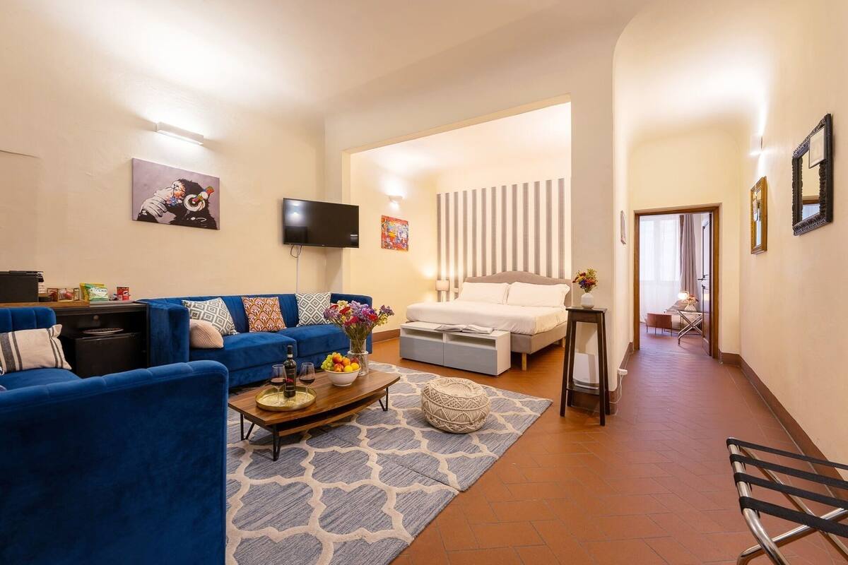 A modern living room with a blue sofa, a coffee table with a vase of flowers, and a bed with a striped headboard.