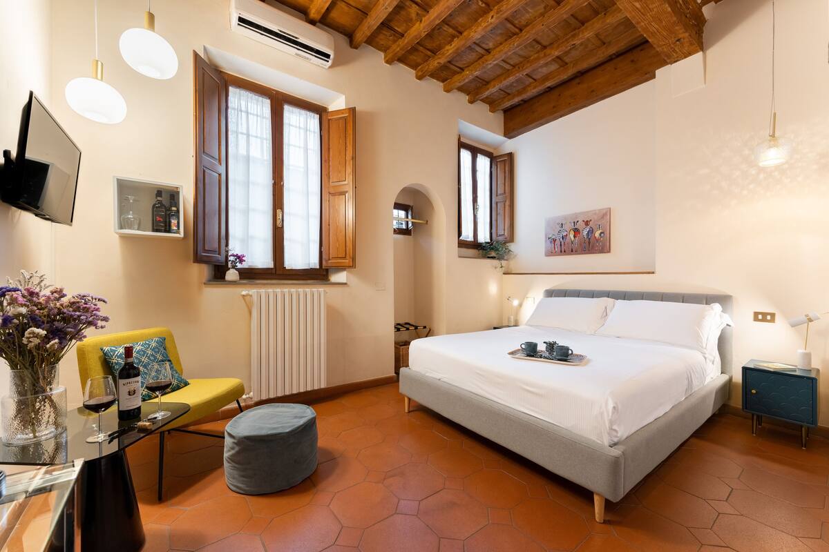 A bedroom with a bed, a small table with a vase and wine glasses, a chair, and a window with wooden shutters.