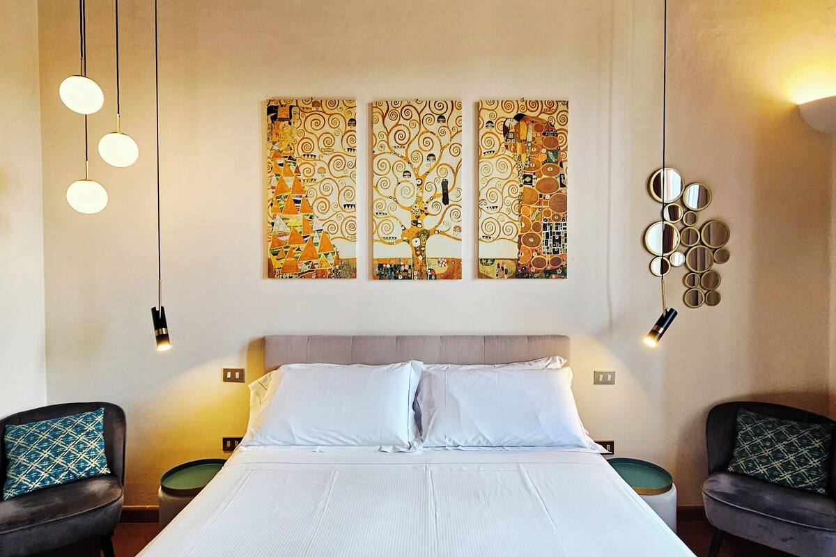 A hotel room with a wooden ceiling, a large bed with white linens, two black chairs with patterned cushions, and a wall with a large, colorful abstract painting.