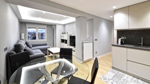 A modern living room with a glass dining table, grey sofa, and a view of the city through large windows.