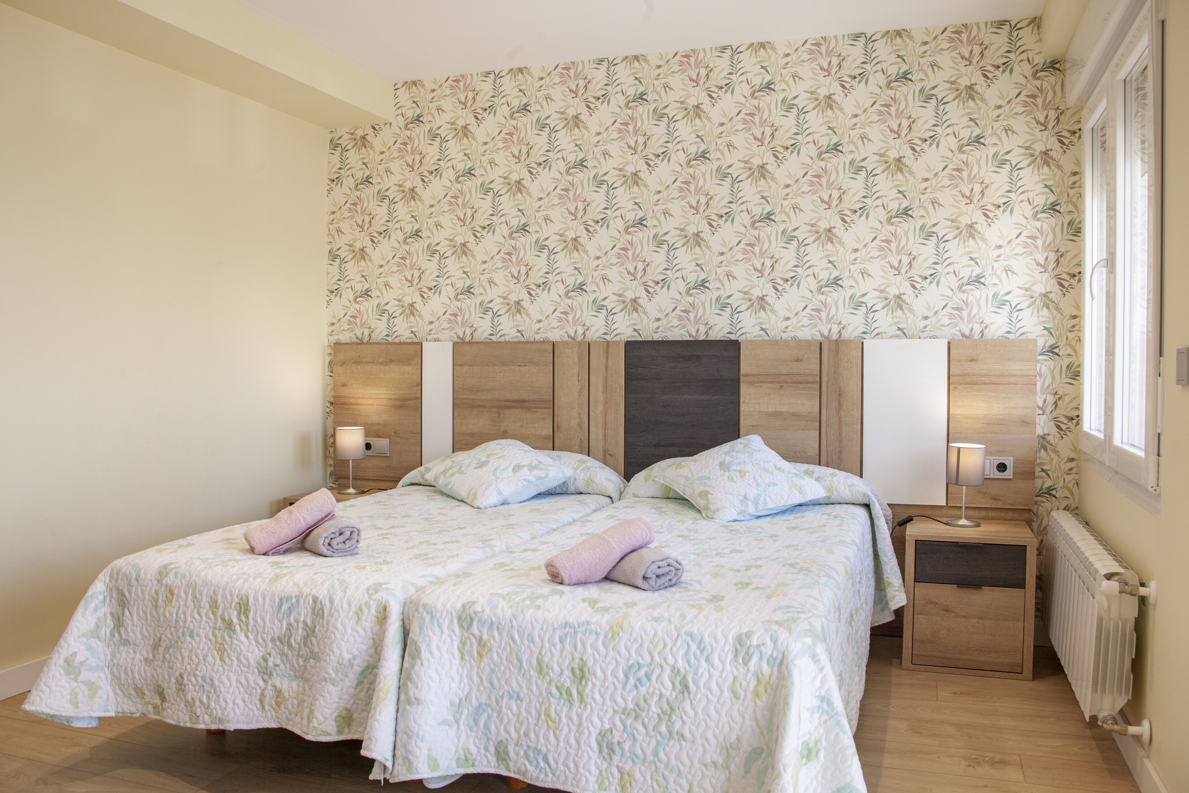 Una cama doble con ropa de cama estampada con motivos florales, cabecera de madera, lámparas de noche y un radiador.