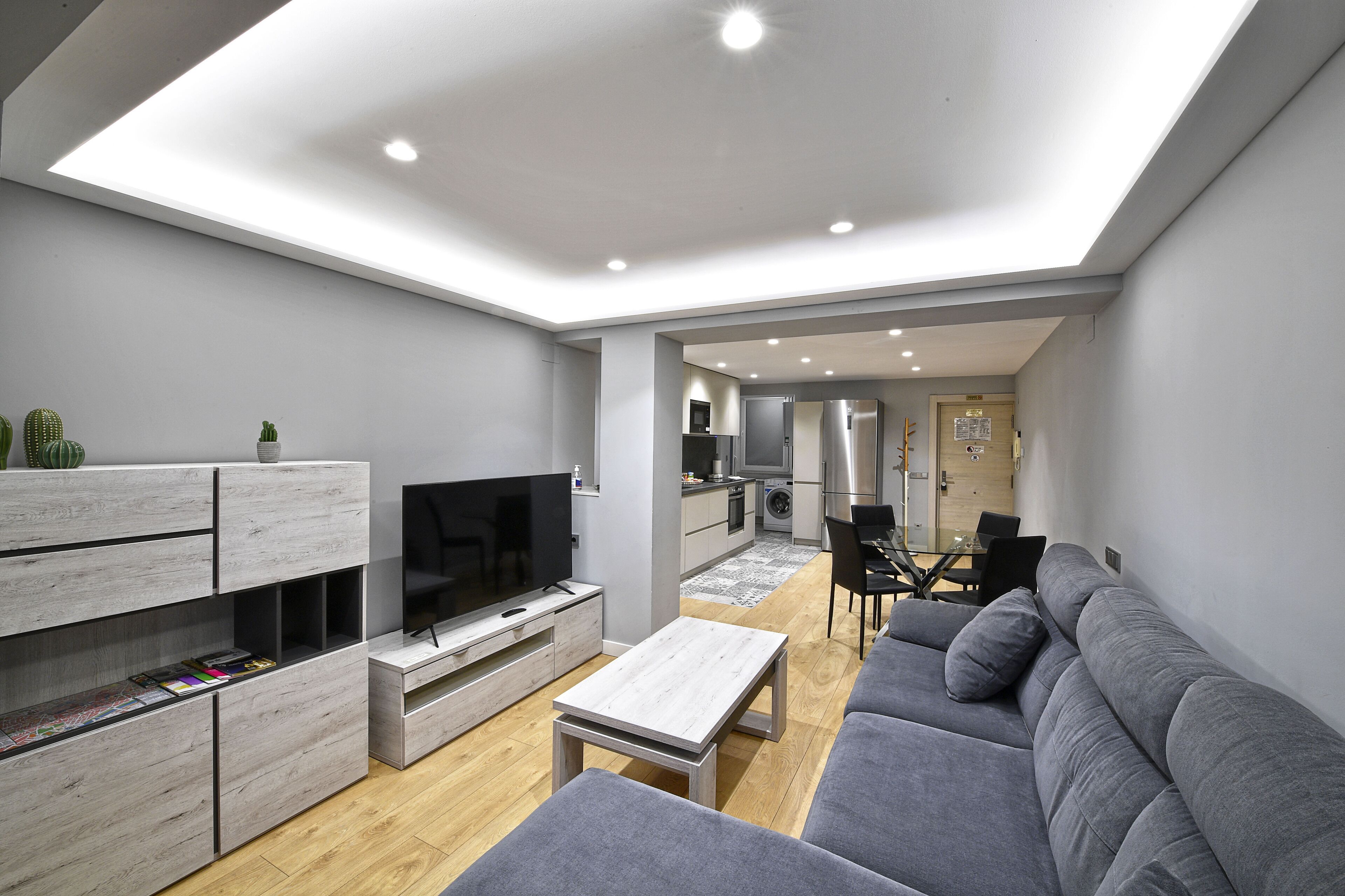 Een moderne woonkamer met een flatscreen televisie, een grijze bank, een houten salontafel en inbouwkasten.