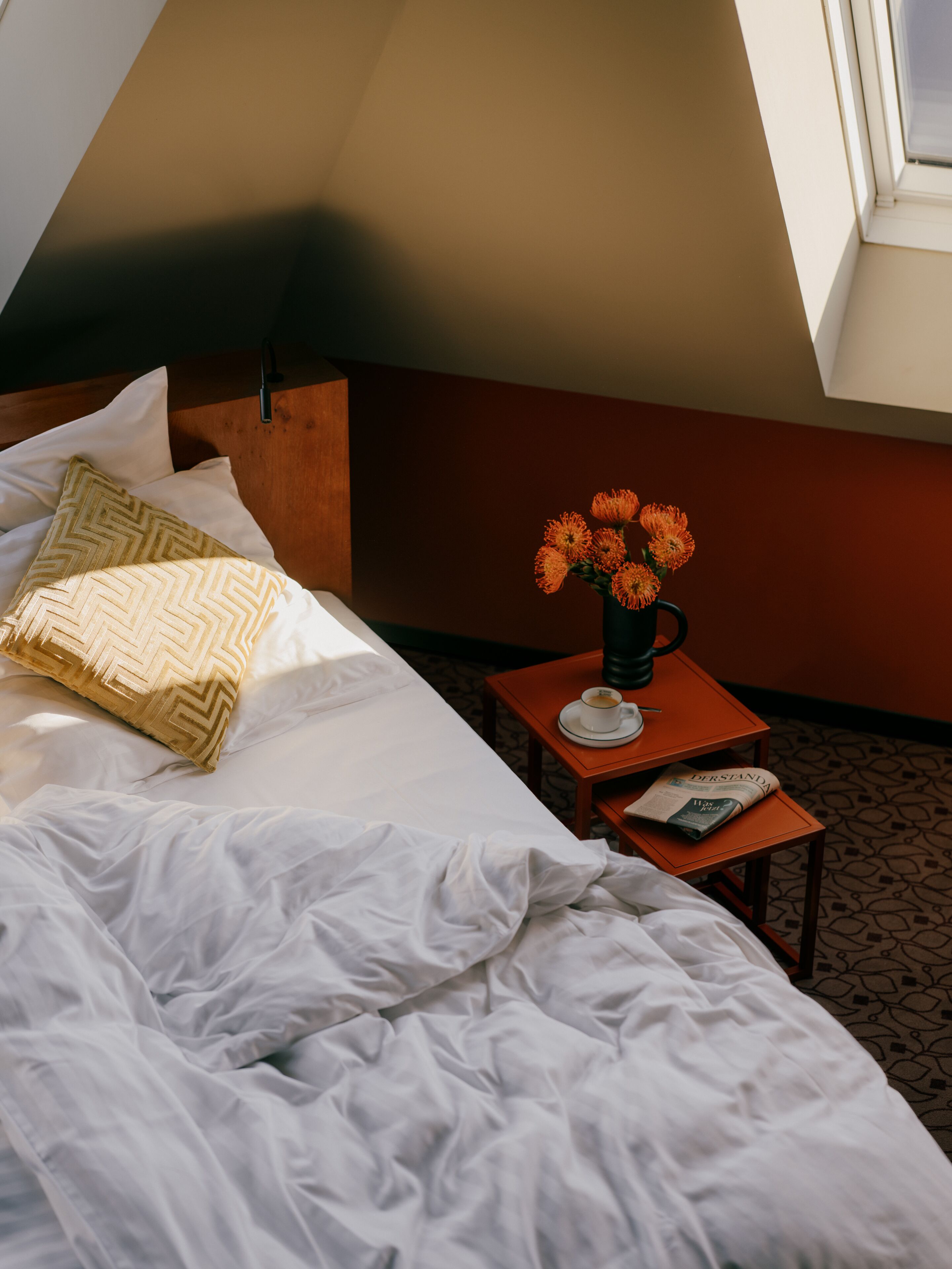 A hotel room with a bed, a small table with a cup of coffee, and a vase of flowers.