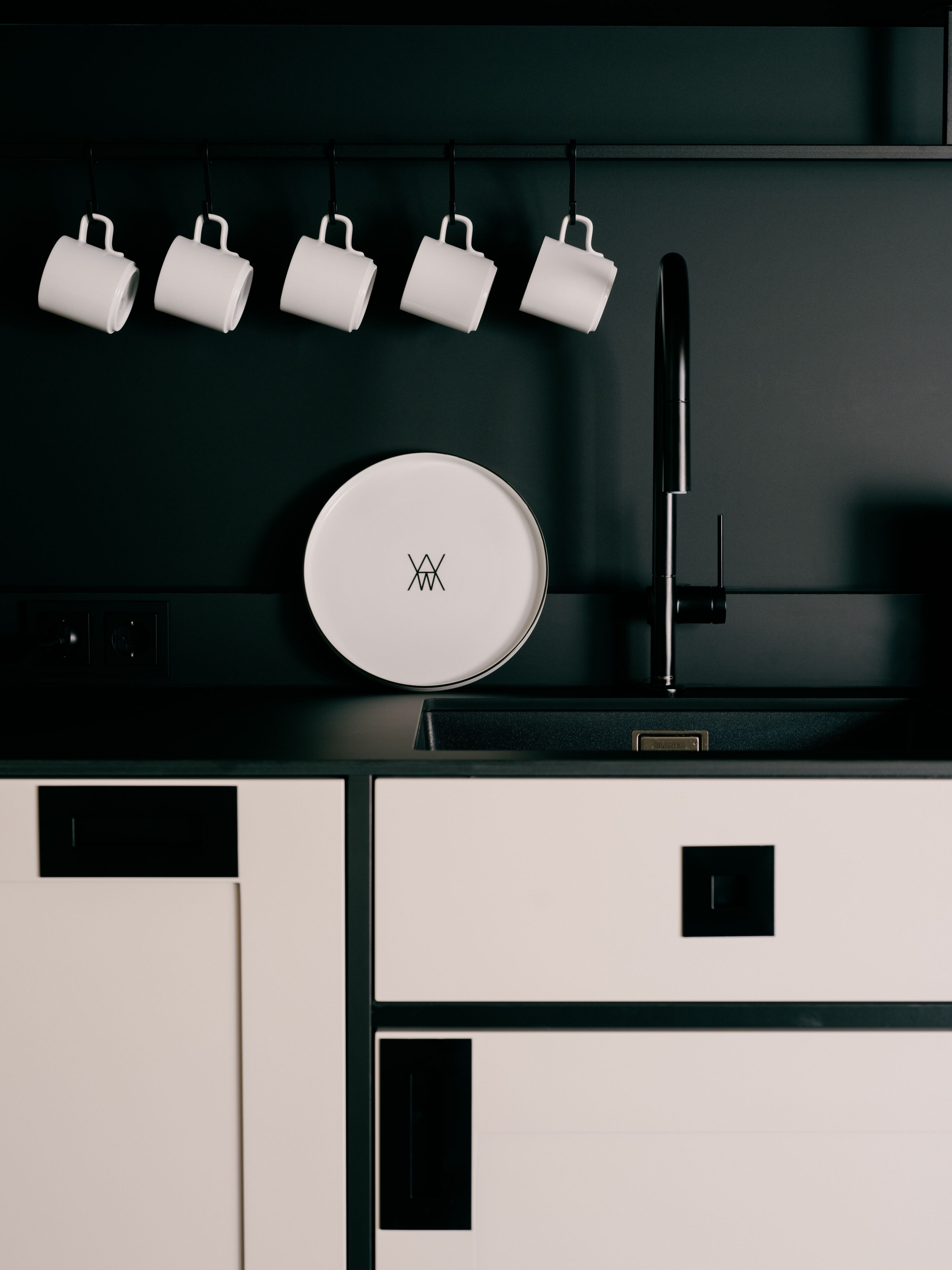 Una cocina con diseño moderno, que incluye un electrodoméstico blanco circular con un logo, un grifo negro y cinco tazas blancas colgadas en un soporte negro.