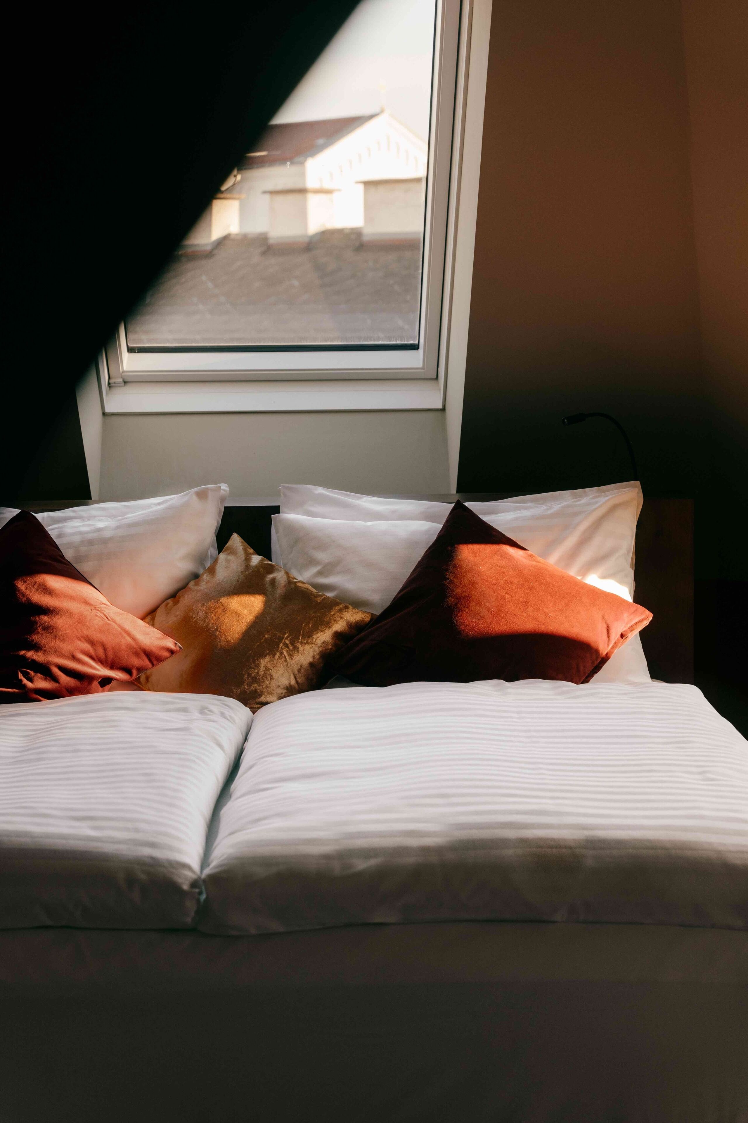 Ein Bett mit weißen Leinentüchern und mehreren Kissen, darunter eines in Braun, in einem Zimmer mit einem Fenster.