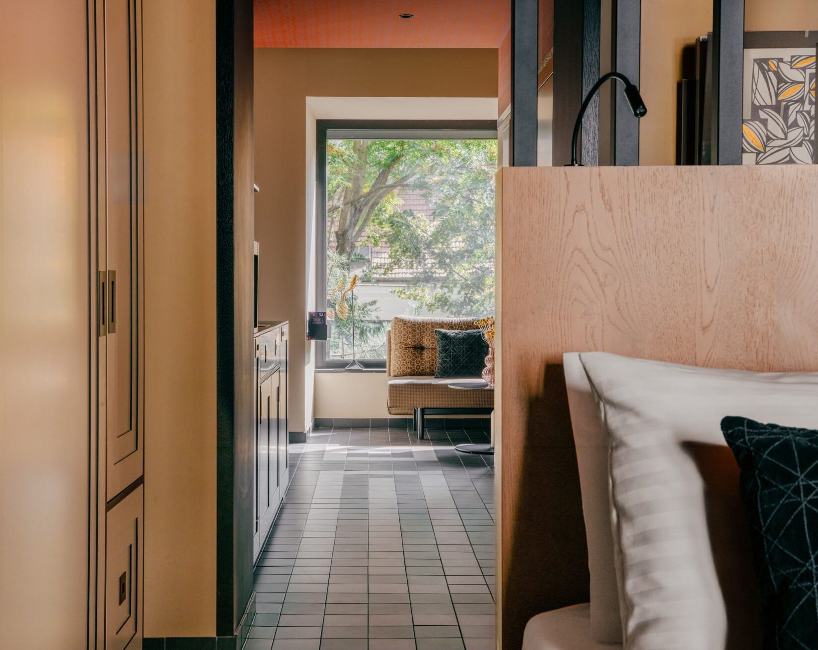 A modern hotel room with a view of trees through a large window, a sofa, and a wooden cabinet.