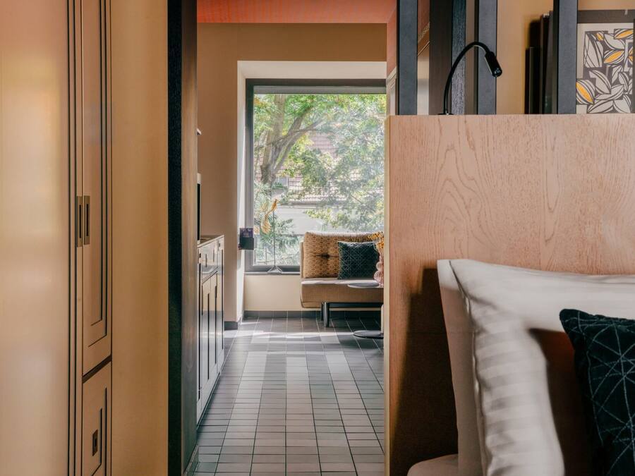 A modern hotel room with a view of trees through a large window, a sofa, and a wooden cabinet.