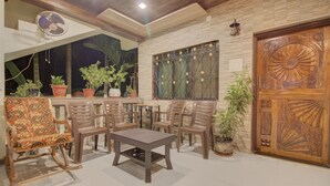 Uma área externa com mesas e cadeiras, rodeada por vasos de plantas e uma porta de madeira decorativa.