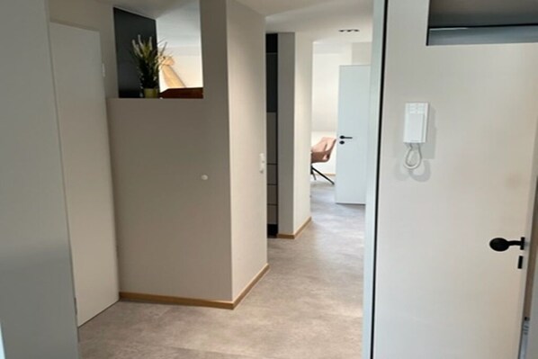 Un couloir avec une porte sur la droite, une petite plante sur la gauche, et on aperçoit une pièce au fond, avec une chaise et une table.