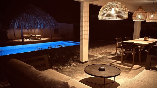 A poolside area with a thatched umbrella, outdoor seating, and hanging pendant lights. - Belnem Garden Residence (Kralendijk)