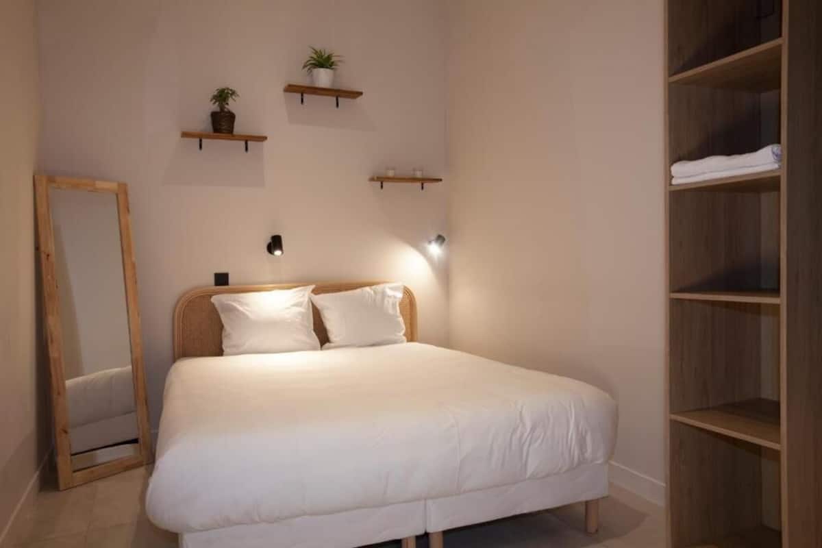 A neatly arranged bedroom with a bed, a mirror, and shelves with plants and towels.