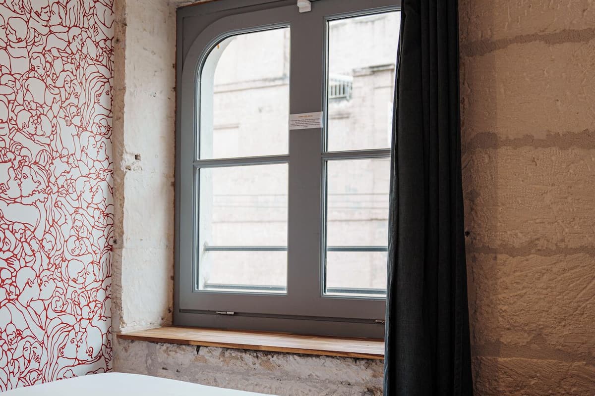 A room with a large bed, a window with a view of buildings, and a textured wall with a red line pattern.