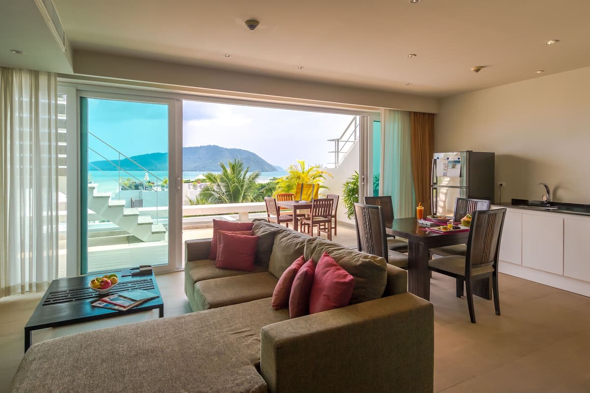 A modern living room with a sofa, a dining table, and chairs. There is a balcony with a view of the sea and mountains.