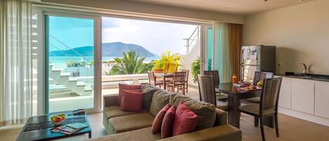A modern living room with a sofa, a dining table, and chairs. There is a balcony with a view of the sea and mountains.