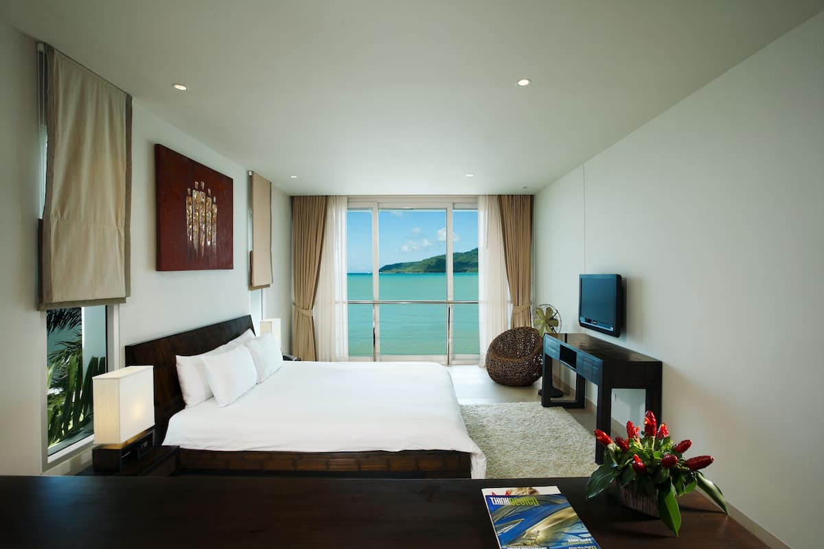 A hotel room with a large bed, a TV, a view of the sea, and a window with curtains.