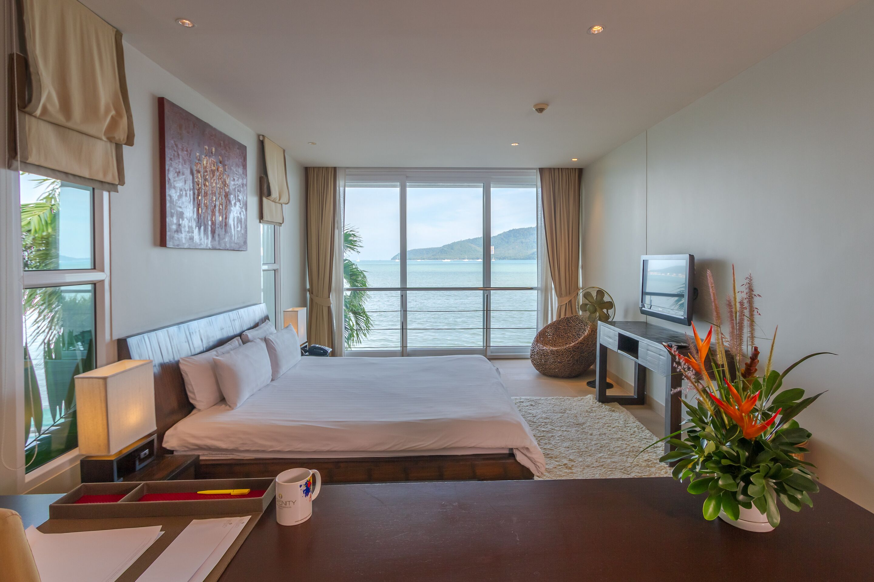 A hotel room with a large bed, a TV, a desk, and a view of the sea.
