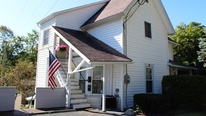 A two-story white house with a dark roof, a front porch with steps, and an American flag. - Cooperstown Baseball Rentals - Team Basemen (Oneonta)