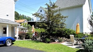 A residential driveway with a blue car parked on the left, a white car on the right, and a small patio area with outdoor furniture and umbrellas. - Cooperstown Baseball Rentals - Team Basemen (Oneonta)