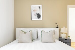 A neatly made bed with white linens and two pillows, a bedside table with a lamp and a small plant, and a framed abstract painting on the wall. - Luxurious Villa in Shepherd's Bush (London)