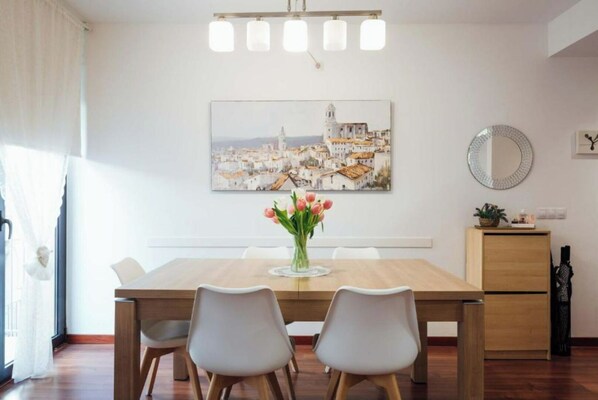 A dining area with a wooden table, two chairs, a vase of flowers, and a painting of a cityscape on the wall.